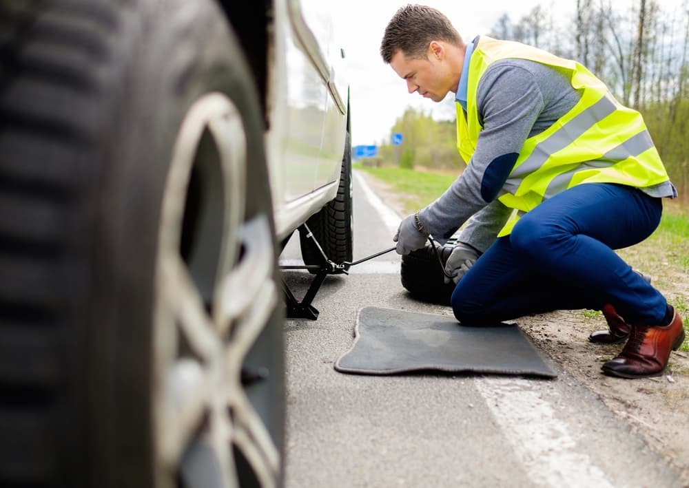 A man wearing protective gear uses tools to change a tire at the side of the road.