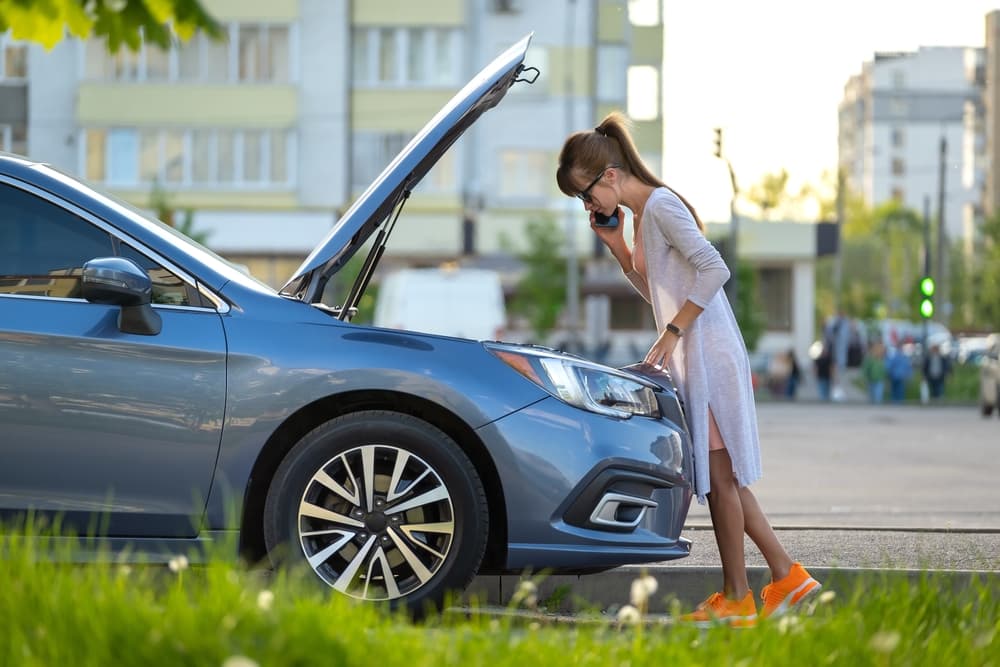 A woman looks at her car engine with the hood open while talking on the phone.